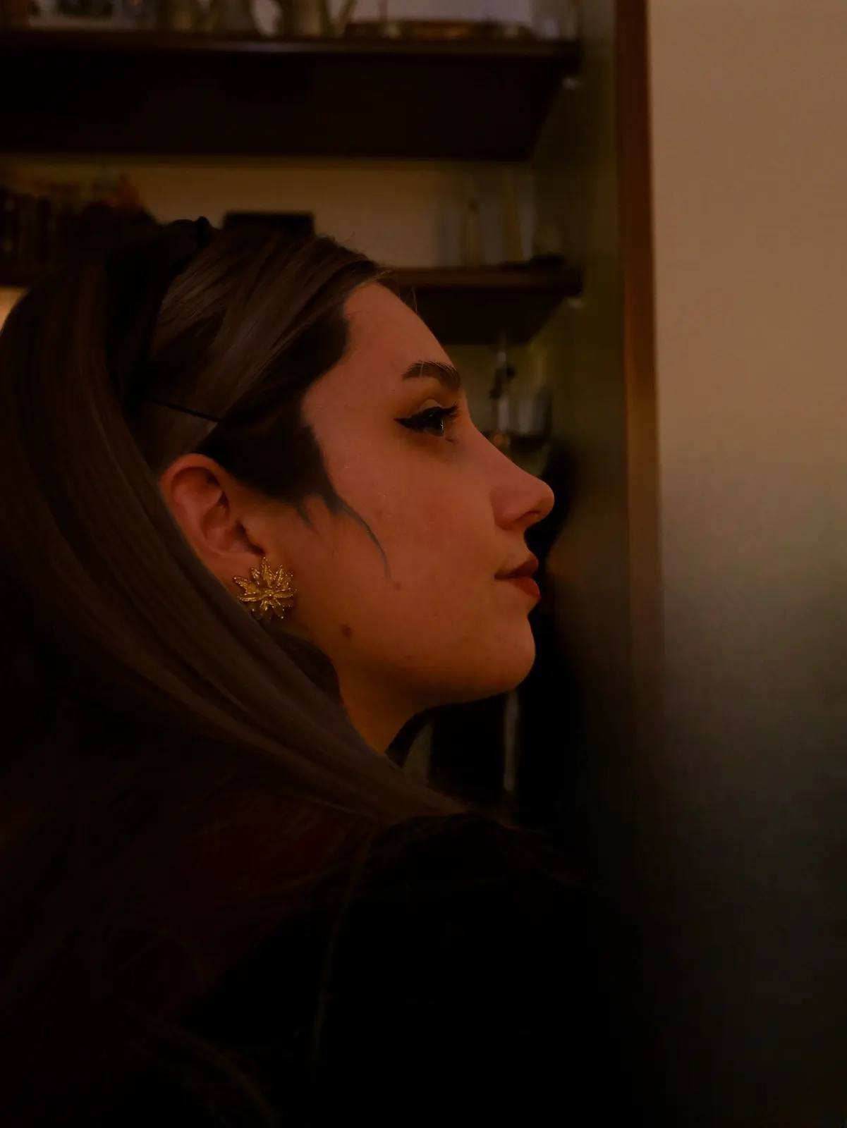 Side-profile portrait of a woman with long brown hair, winged eyeliner, and a gold floral earring in warm, dim lighting.